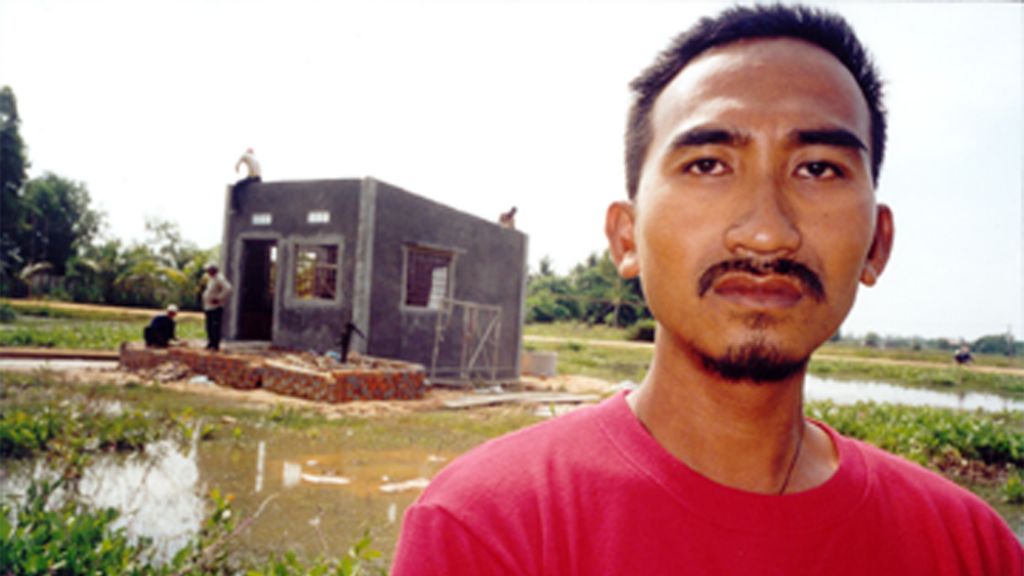 A man stands in front of a muddy tropical field, in front of a one-room cement structure