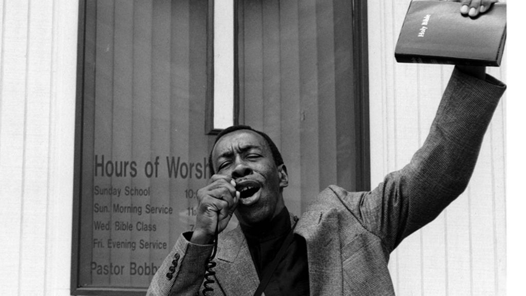 A man preaches on a street corner in front of a large cross on a window, holding a bullhorn and a Bible