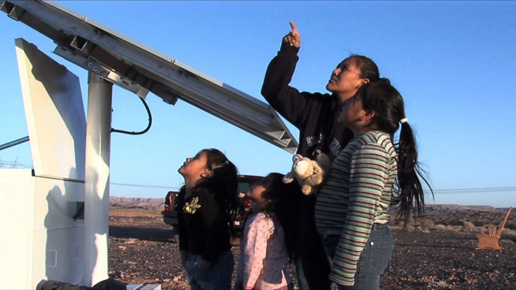 A woman and three kids look on as a solar panel is set up