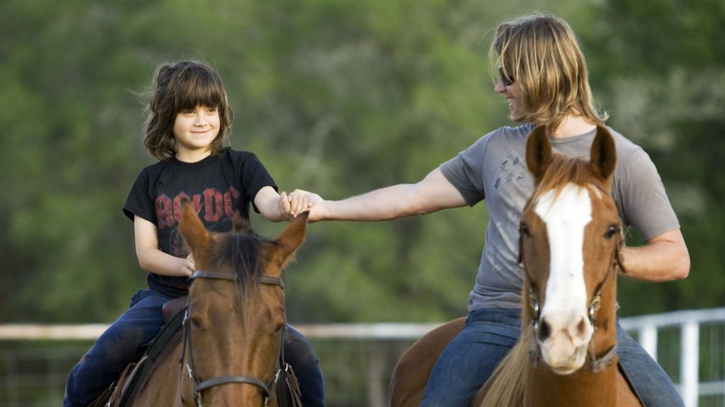 A child and an adult hold hands while riding two separate horses.