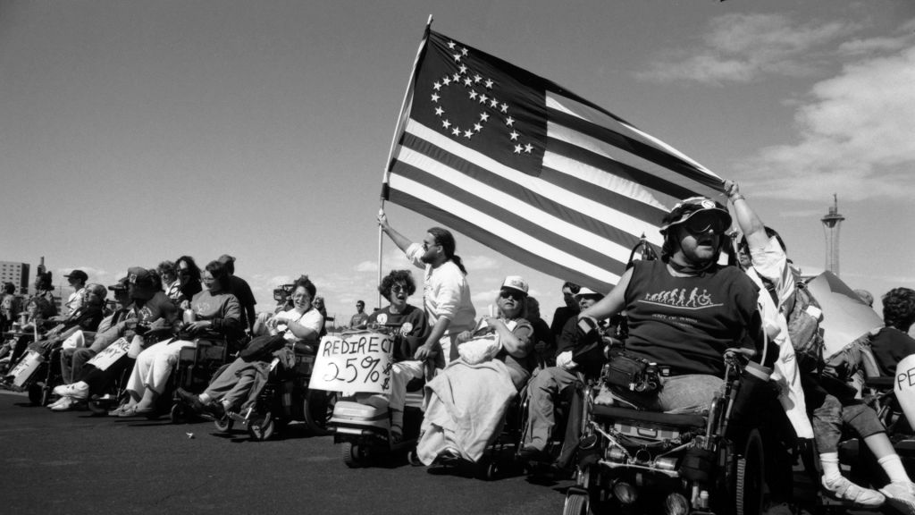 Disability rights activists assemble under an American flag with its stars arranged in the shape of a wheelchair.