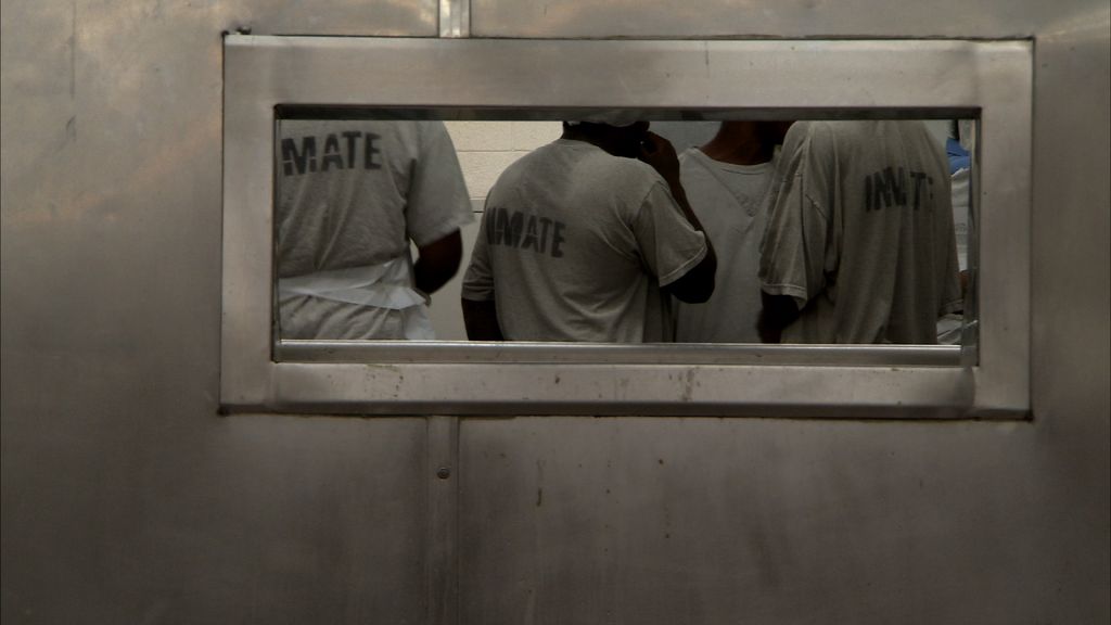 Inmates gather in a holding cell, seen through a viewing slot in a secure metal door.