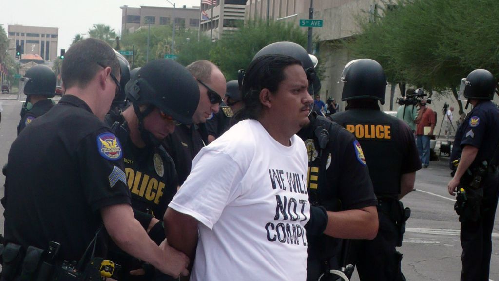 Immigration activist wearing "We Will Not Comply" T-shirt arrested by a group of police.