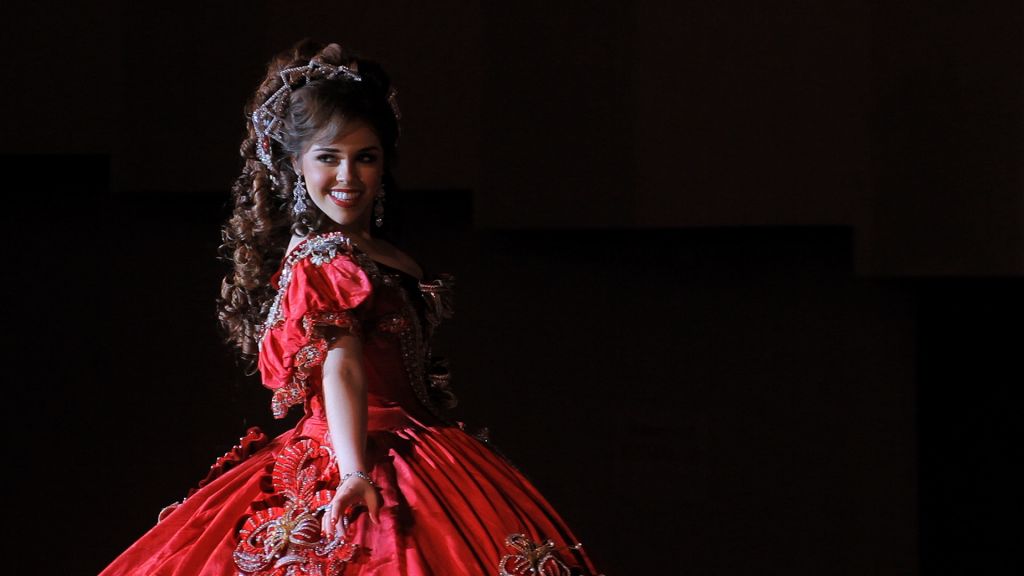 Young woman in elaborate red dress poses on stage during debutante ball.