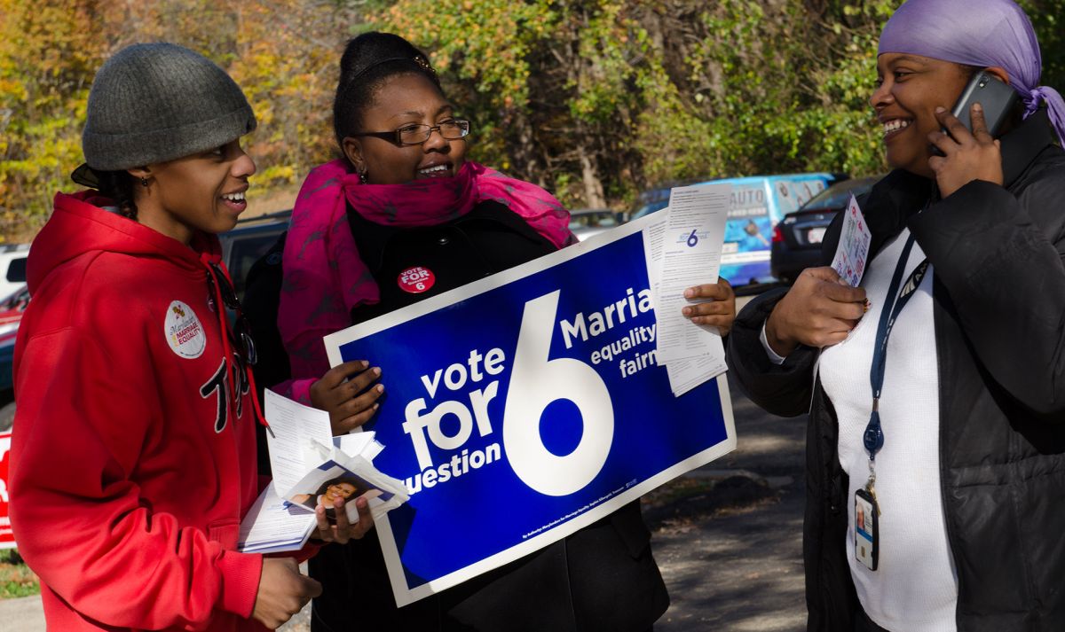 Three people, all with dark skin tone, stand together on Election Day wearing voting stickers. One holds a sign that says "Vote for... marriage equality."