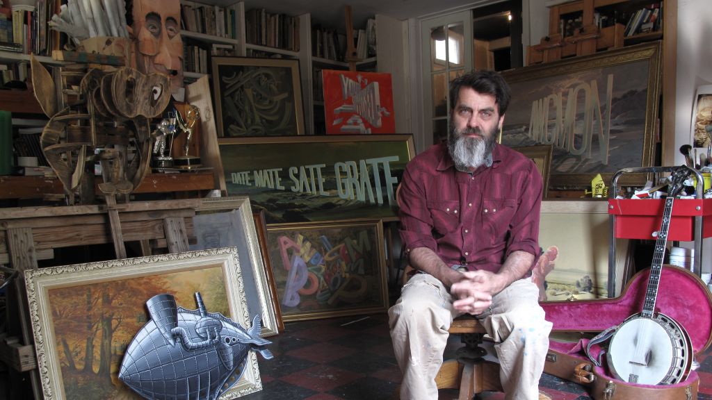 Artist Wayne White poses in his studio with samples of his artwork in the background.