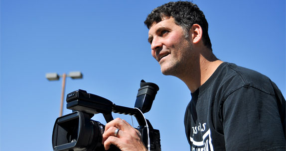 Filmmaker Brad Lichtenstein smiles while framing a shot with a video camera.