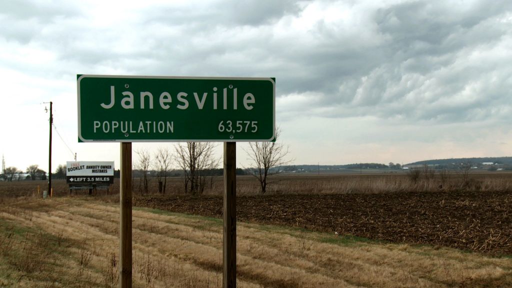 Highway sign in rural countryside reads "Janesville Population 63,575."