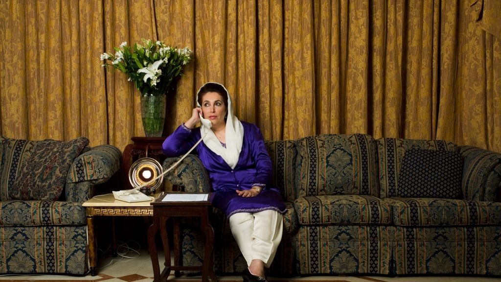 Benazir Bhutto speaks on the telephone while seated on a couch with a gold curtain in the background.