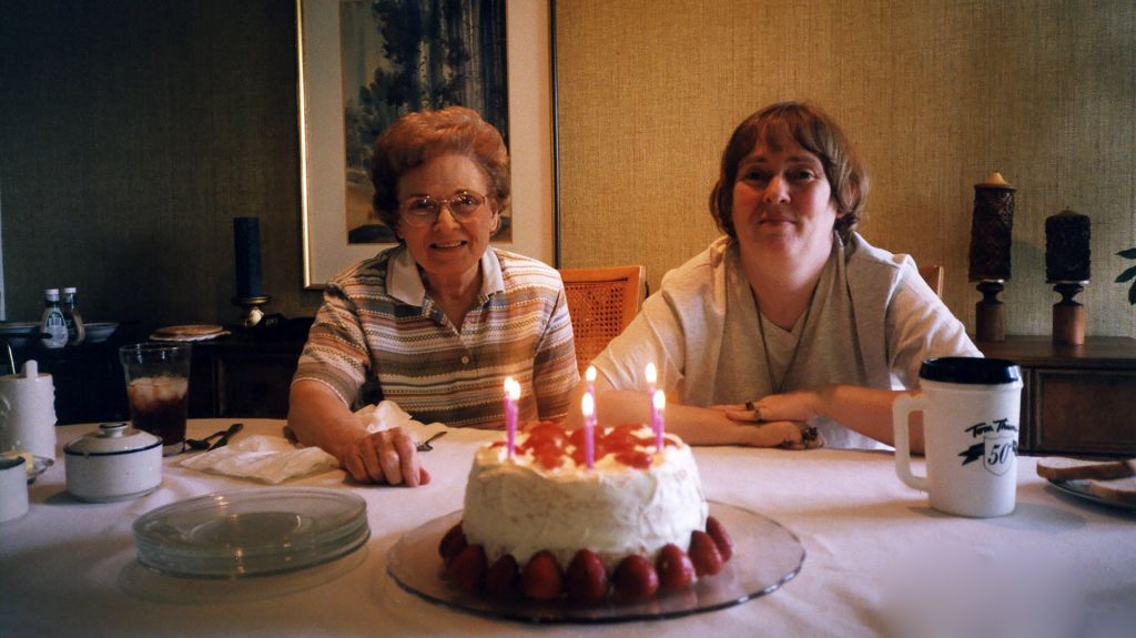 Elderly mother and grown daughter sit behind a birthday cake.