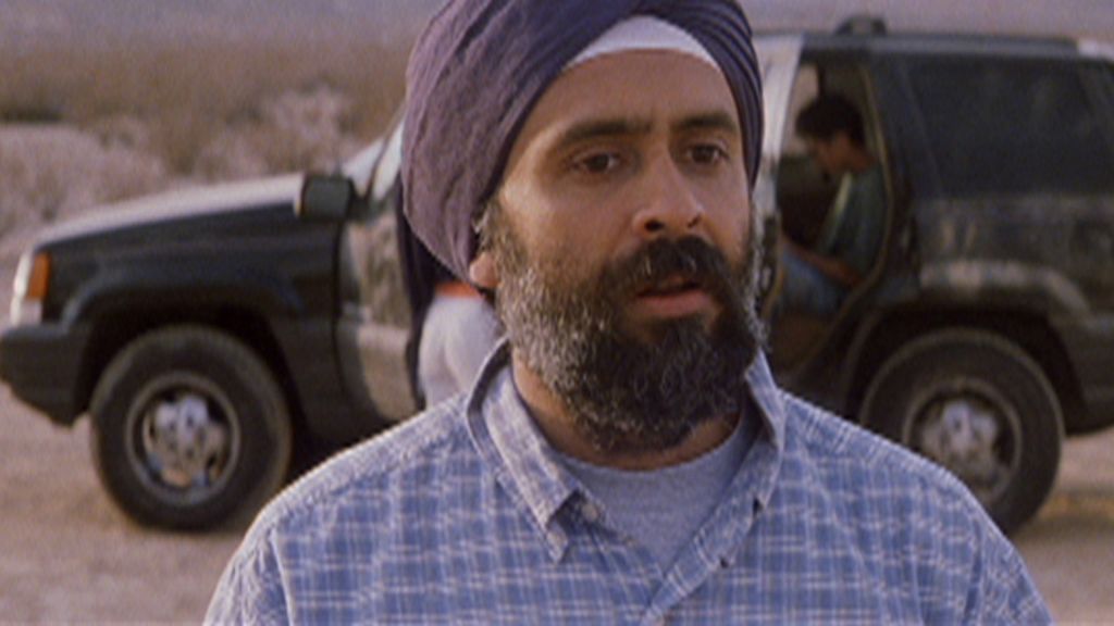 A man with a beard wearing a dark grey turban and a checkered shirt stands in front of an SUV in the desert