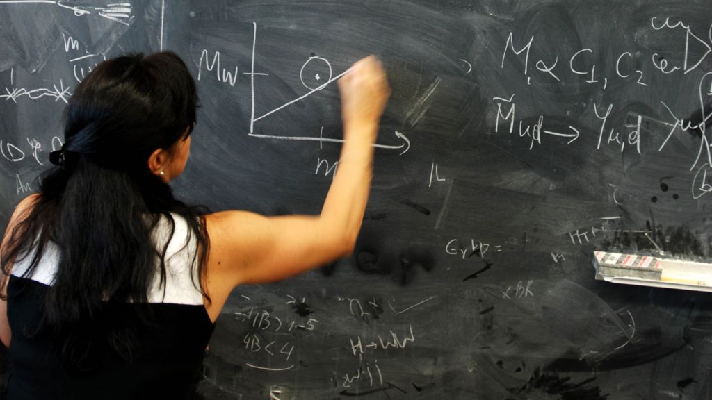 A woman writes a physics formula on a blackboard