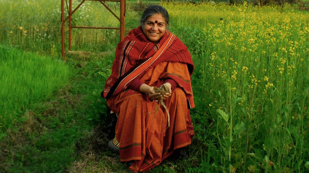 A woman kneels in a field holding a handful of dirt and smiling