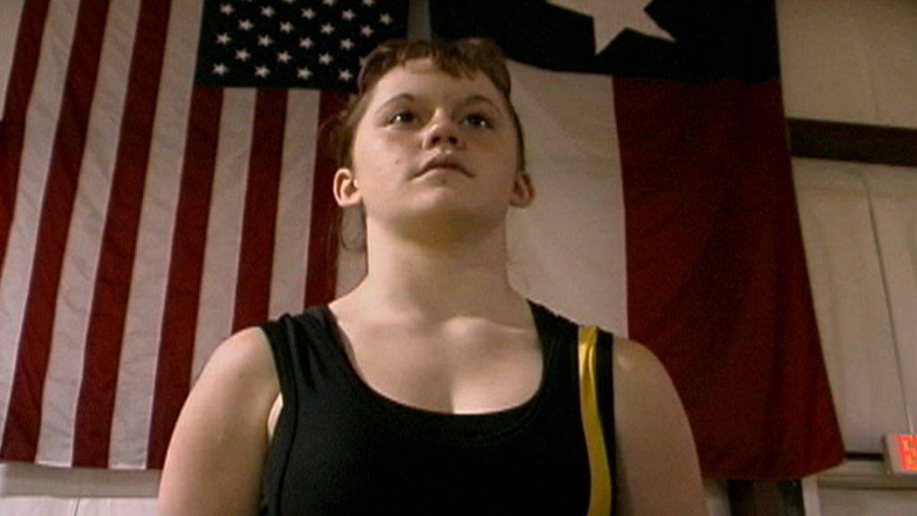 A girl stands in her wrestling uniform in front of large American and Texas flags