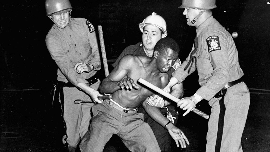 Three white New York State police wearing riot gear and holding nightsticks struggle to subdue a shirtless black man