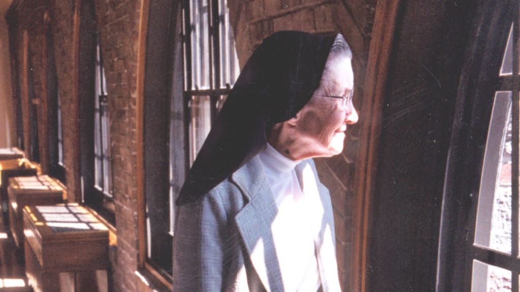 An elderly nun in a habit looks out a window