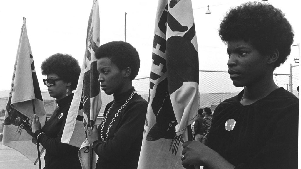 Three women members of the Black Panther Party stand with flags.