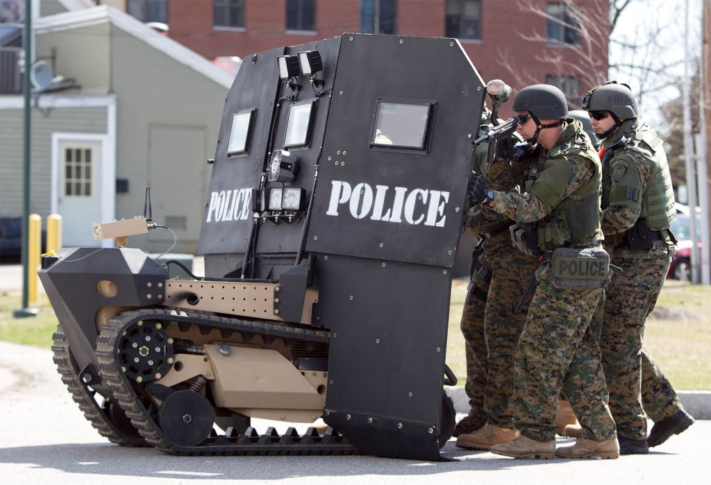 Police in military fatigues huddle behind a SWAT robot