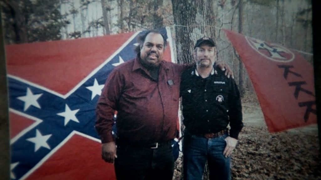 Musician Daryl Davis stands with a member of the Klan next to a confederate flag and a KKK flag