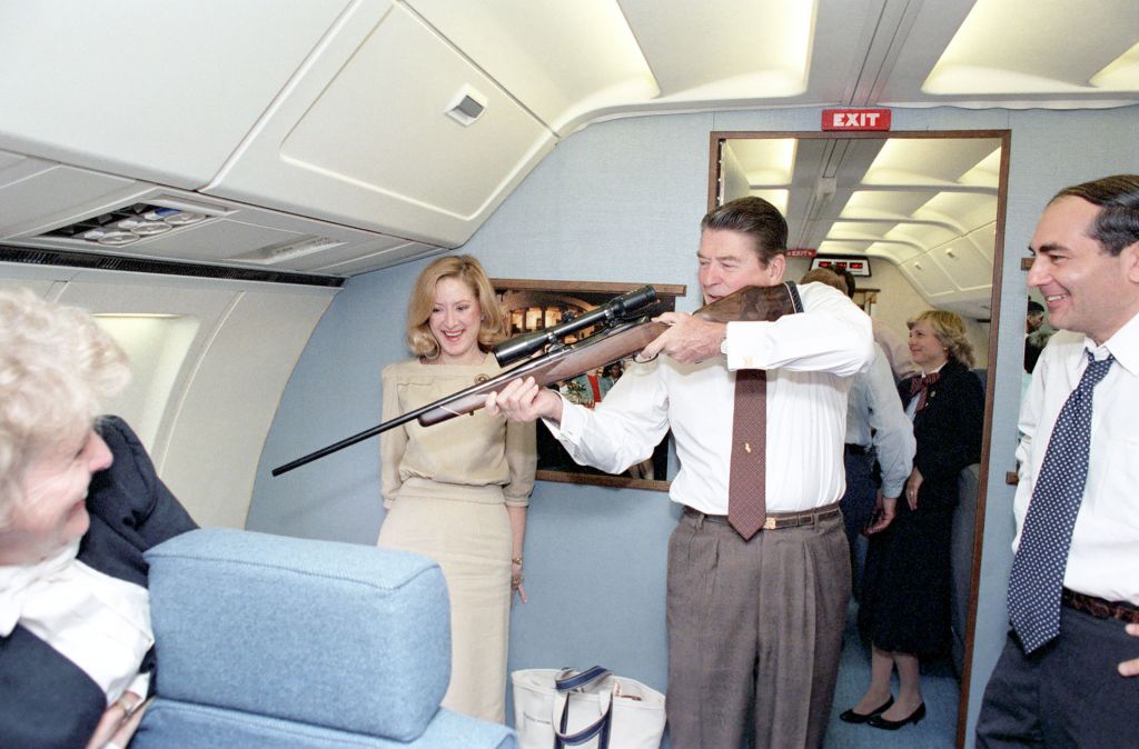 President Reagan aiming a rifle at a window while flying aboard Air Force One during a trip to California,11/23/83. From Shadow World.