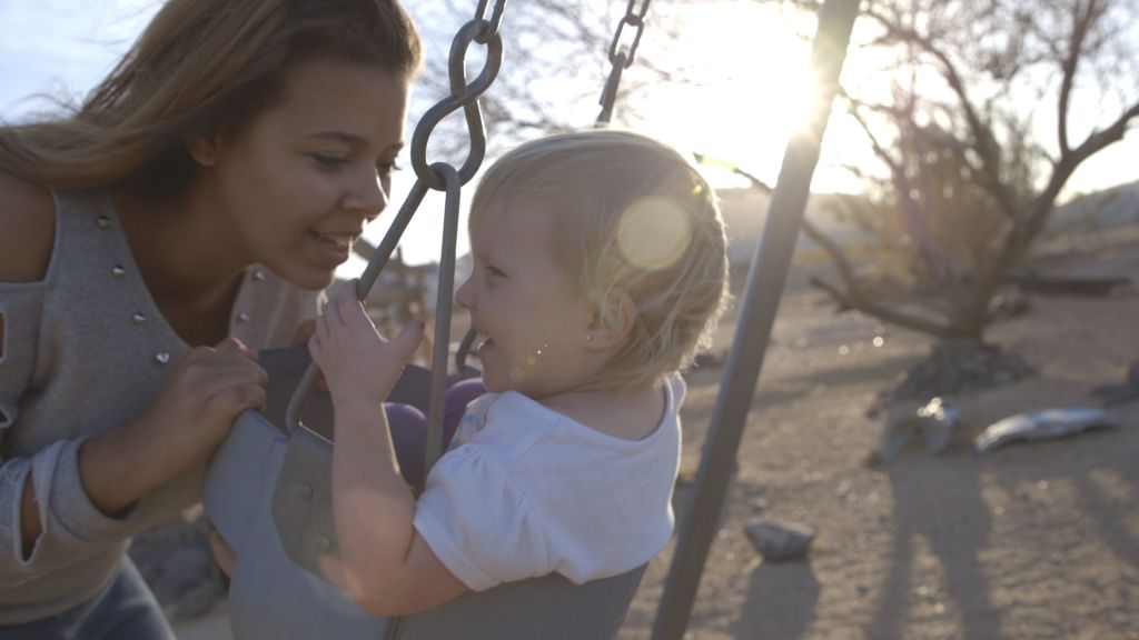 A young woman smiles at a toddler riding a swing set in the desert.