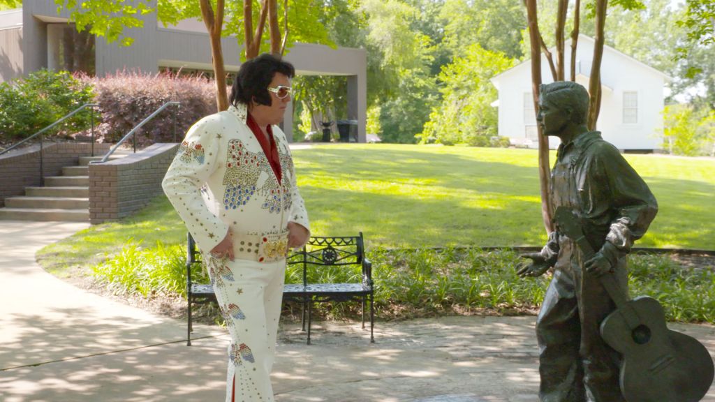 An Elvis impersonator looks at a statue of young Elvis