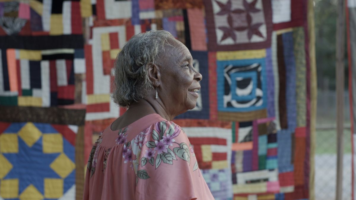 Elderly African American woman standing in front of a colorful quilt.