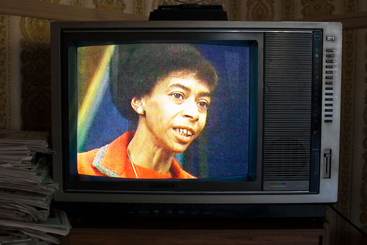 A vintage television set screen shows an image of a Black woman's face. She is wearing an orange blazer.