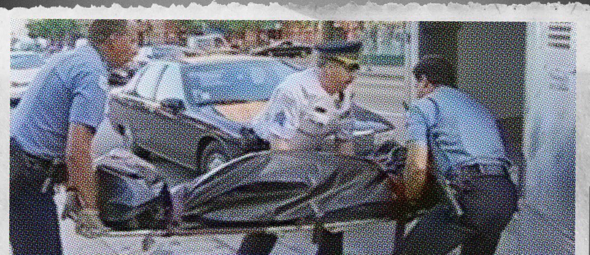 A still from archival news footage showing three men carrying a body bag into the back of an ambulance.