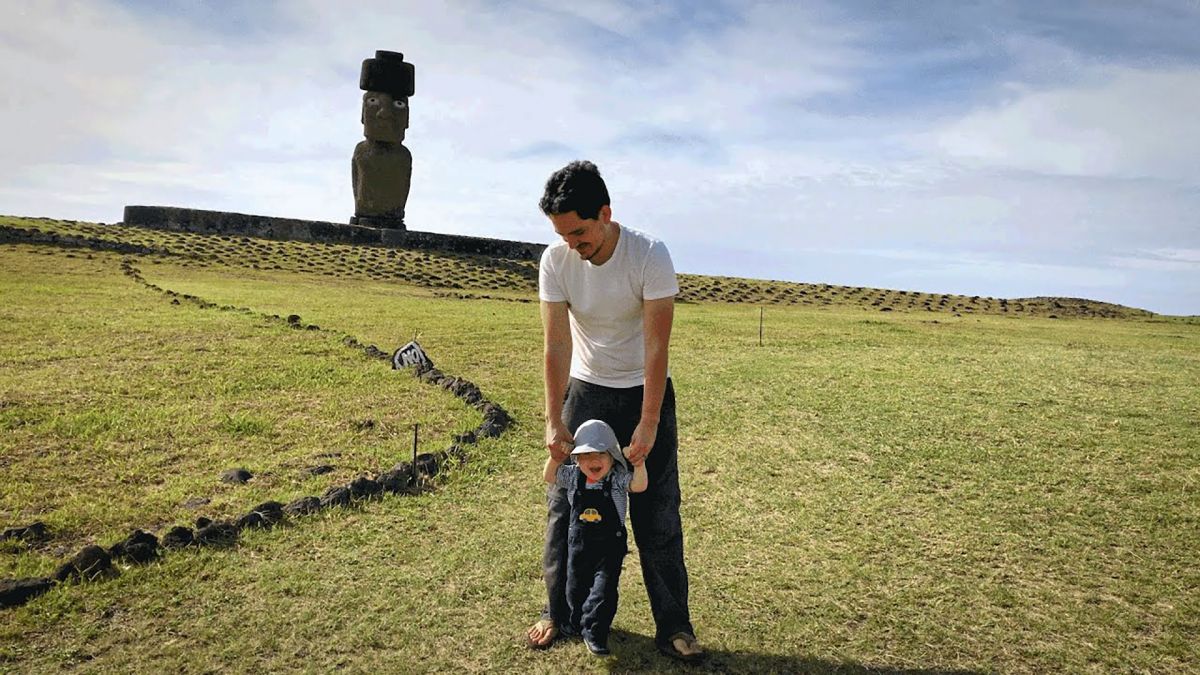 Filmmaker Sergio Rapu walks his infant son down a hillside on Rapa Nui (Easter Island). In the background, a single moai statue stands in the sun.