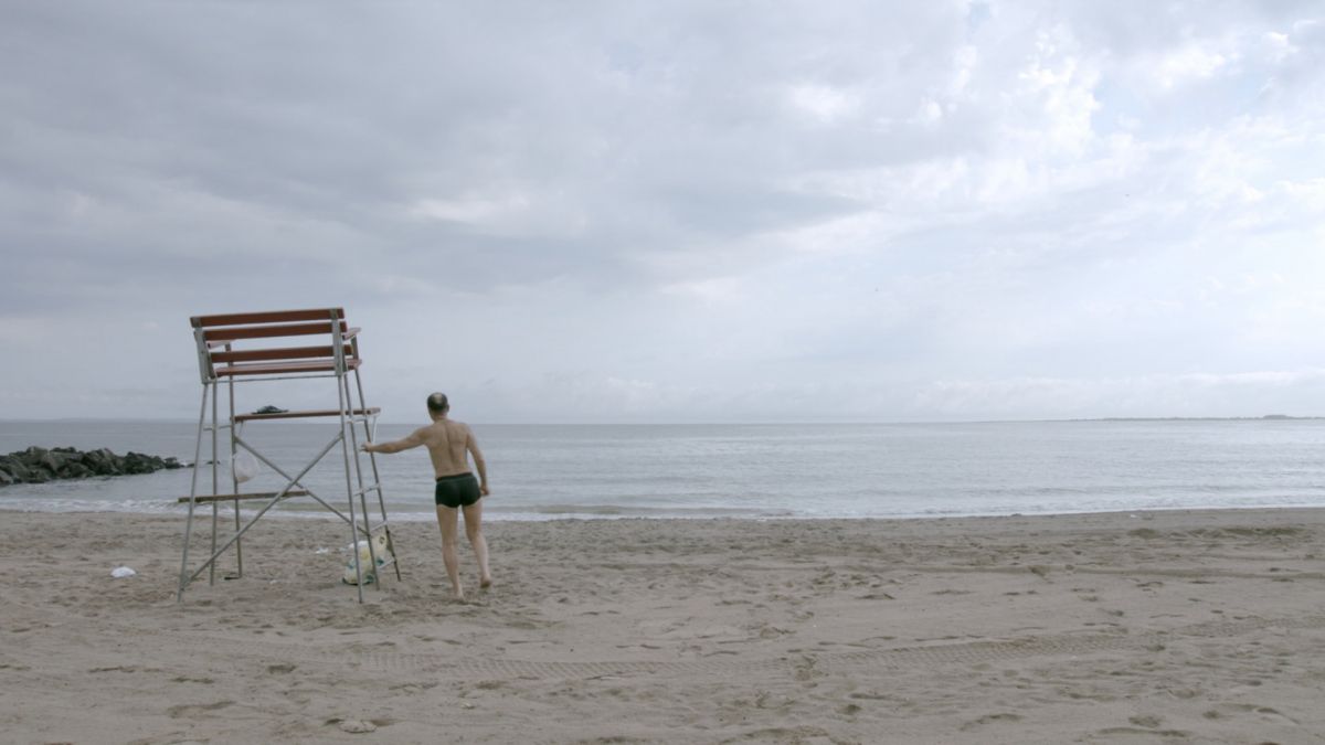 A solitary man with light skin tone stands on a beach on a cloudy day, leaning against a lifeguard stand.