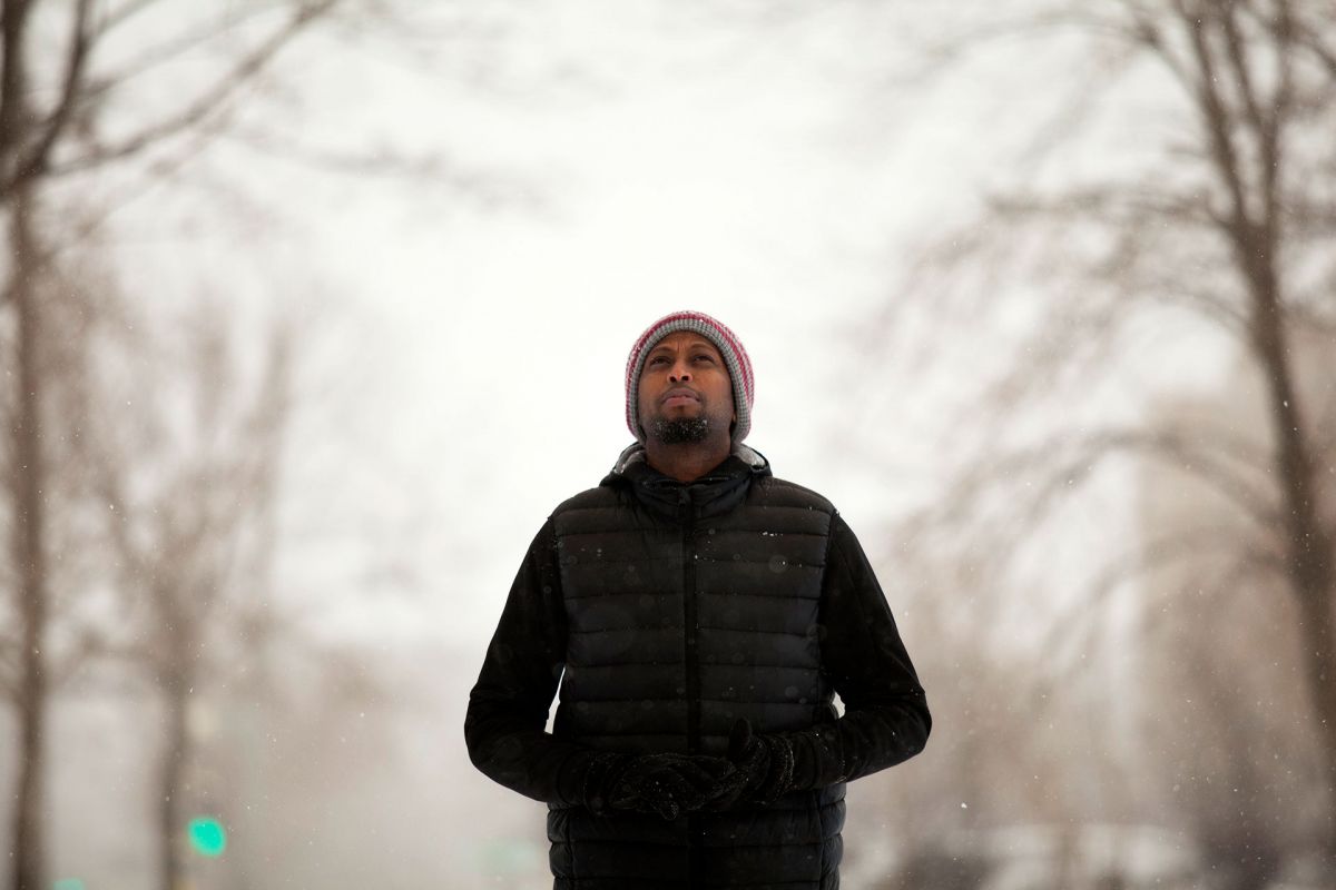 A man with dark skin tone wears a puffer jacket and a beanie as he walks around a snowy park.