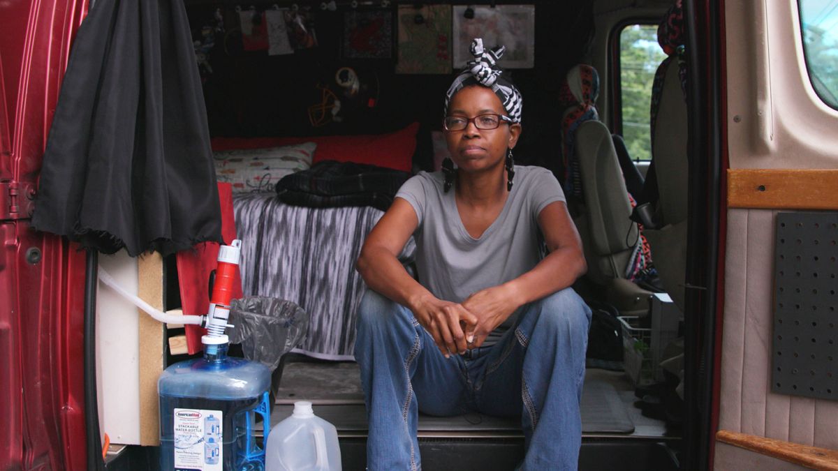 A woman with dark skin tone, wearing a bandana and glasses, sits in the back of a van that's outfitted with a bed and water system.