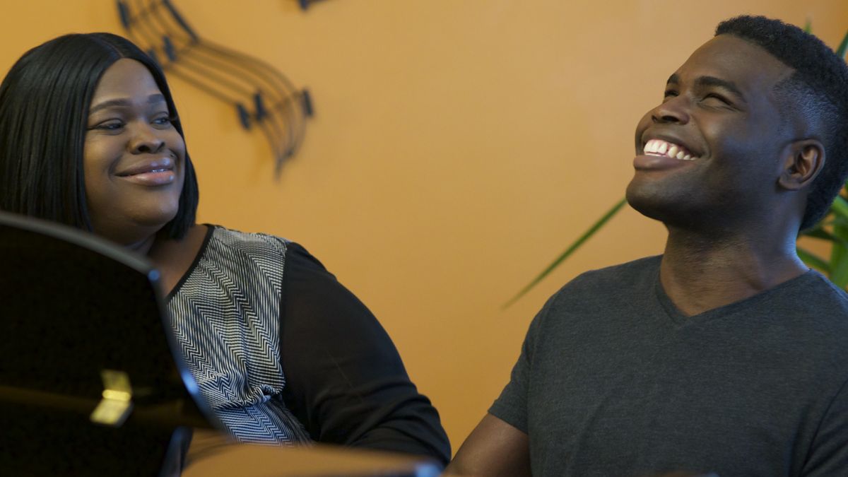 A man and a woman, both with dark skin tone, laugh together while sitting in front of a piano.