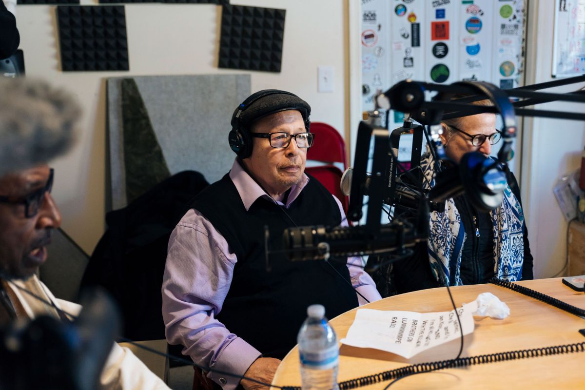 Three older men speak into microphones while recording for a radio program.