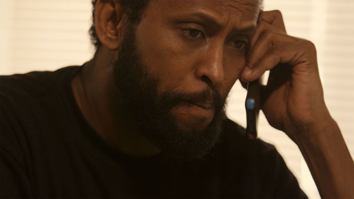 A man with dark skin tone holds a phone to his ear. He looks anxious.