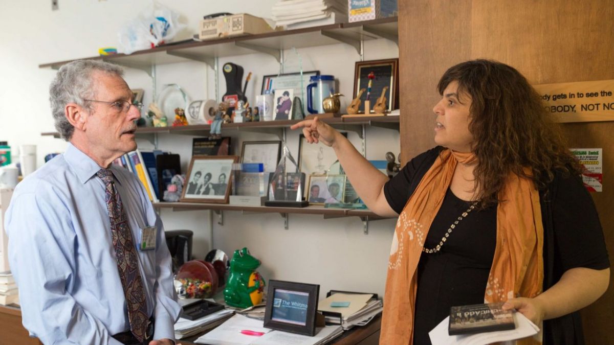 A woman with medium skin tone speaks with a man with light skin tone, gray hair, and glasses inside of an office. She holds up her hand, pointing her finger at him.