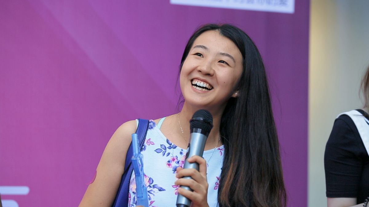 A Chinese woman with long hair speaks into a microphone at an event. She wears a white dress with blue and purple flowers.