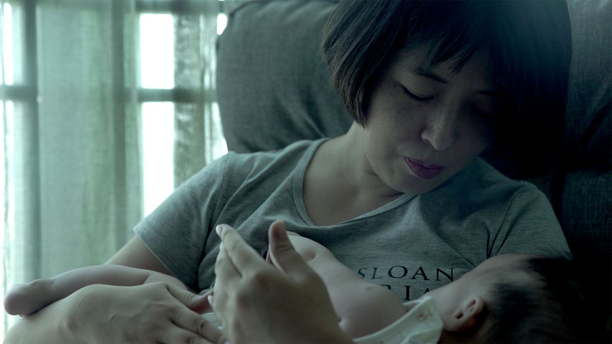 A Chinese woman holds an infant in her arms while sitting in a gray armchair.