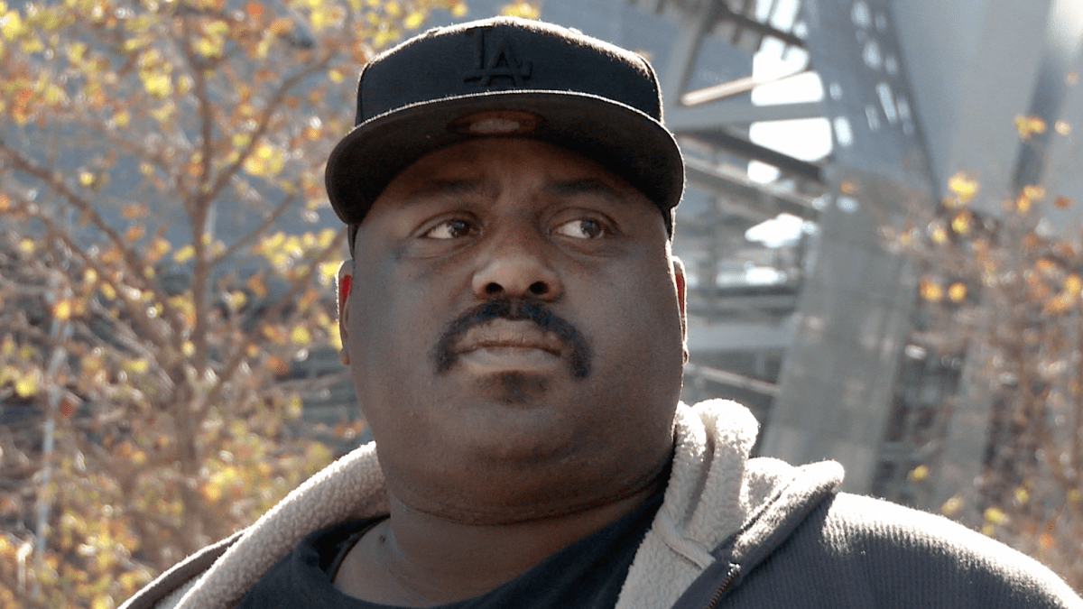 A man with dark skin tone and a mustache, wearing a ball cap, is interviewed for the camera.