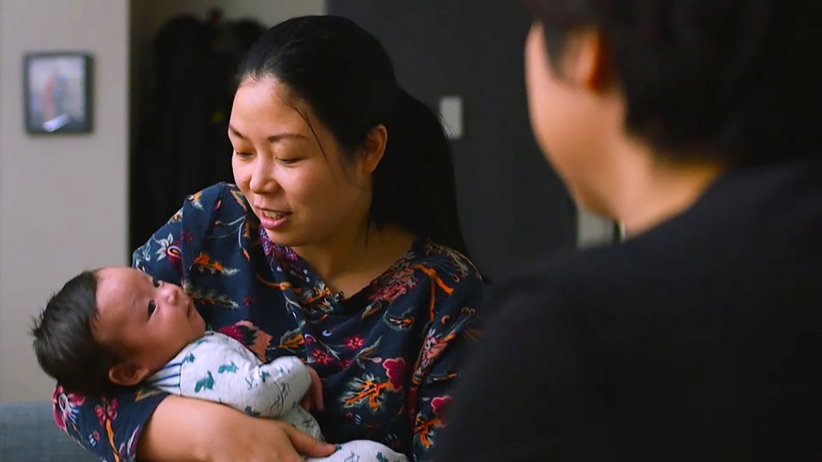 An Asian woman with light skin tone holds her infant child while interviewing her mother for One Child Nation.