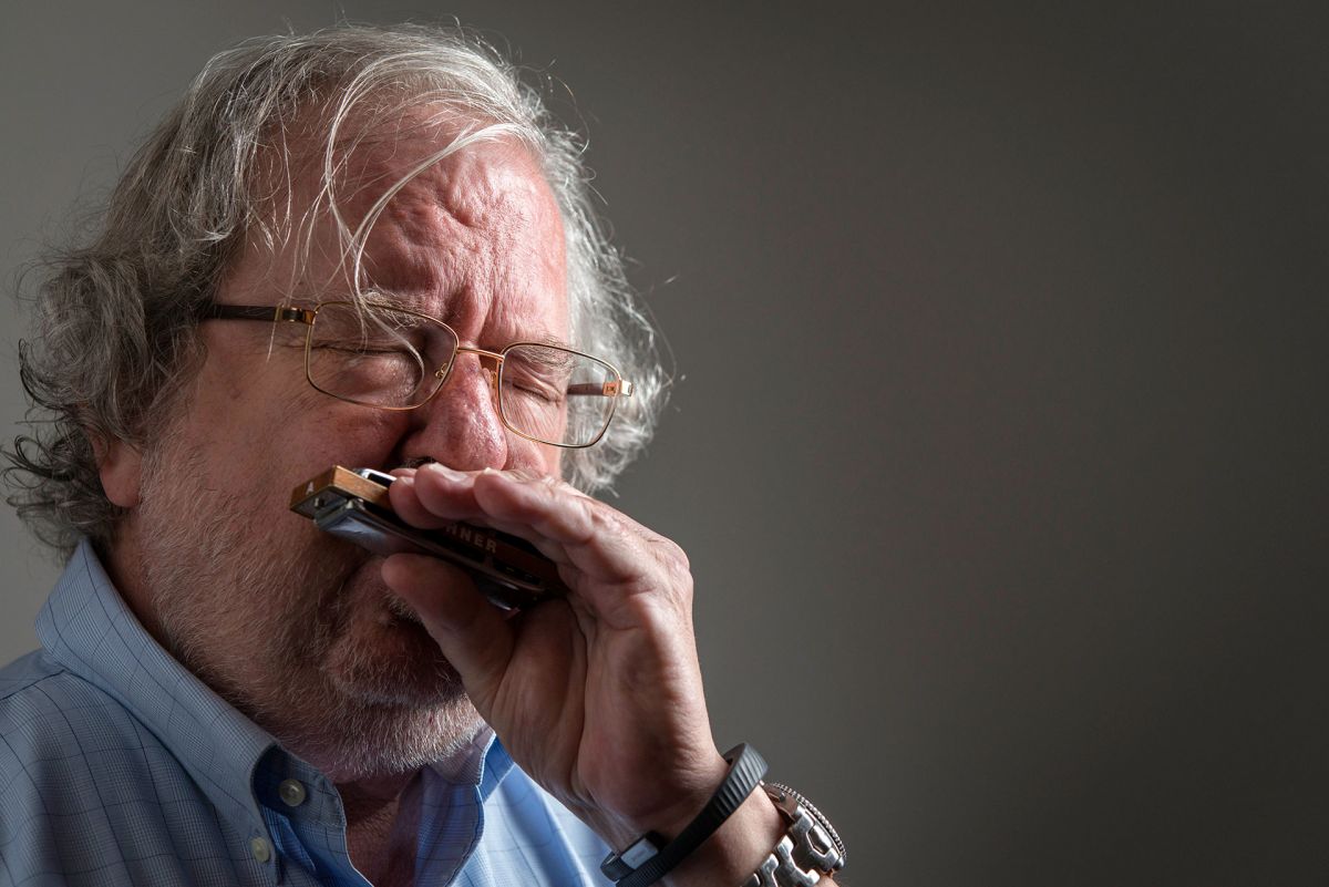 An older man with light skin tone and gray hair, wearing glasses, passionately plays the blues harmonica.