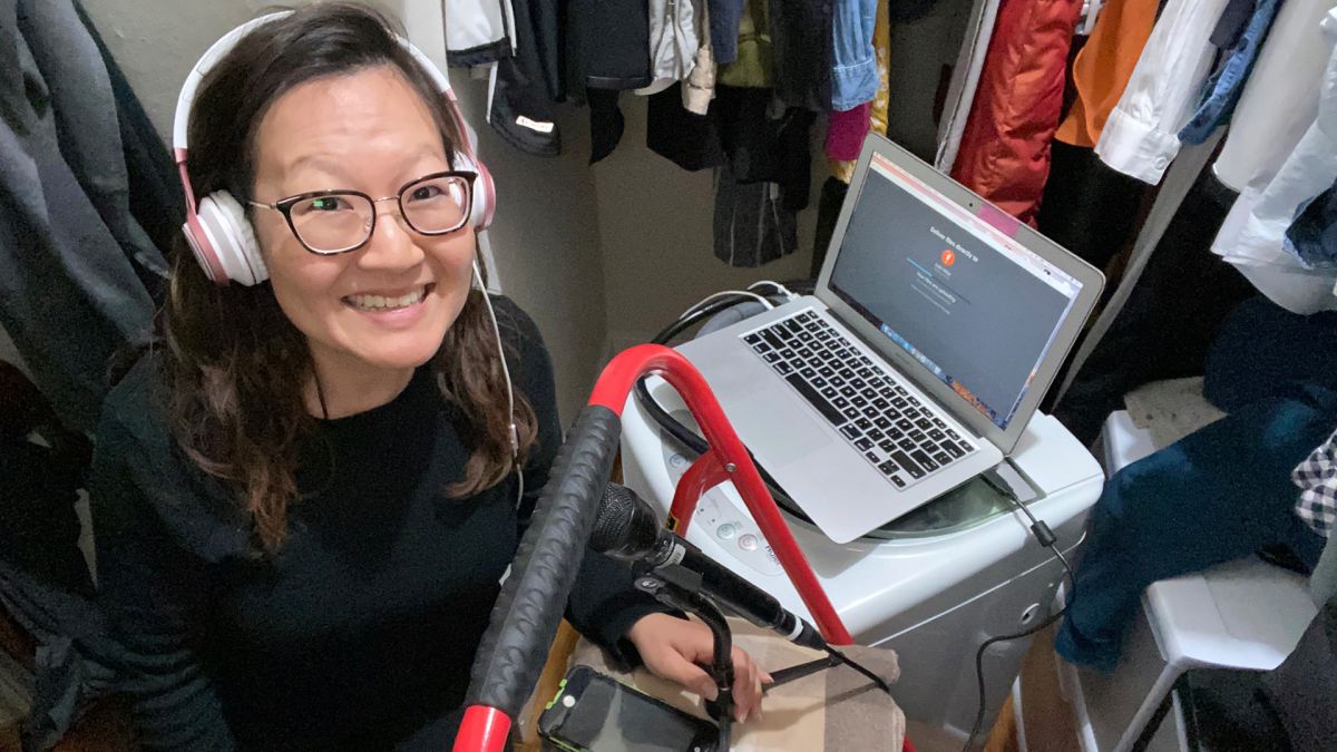 A smiling Asian woman, wearing glasses, uses recording equipment to make a podcast in her closet.