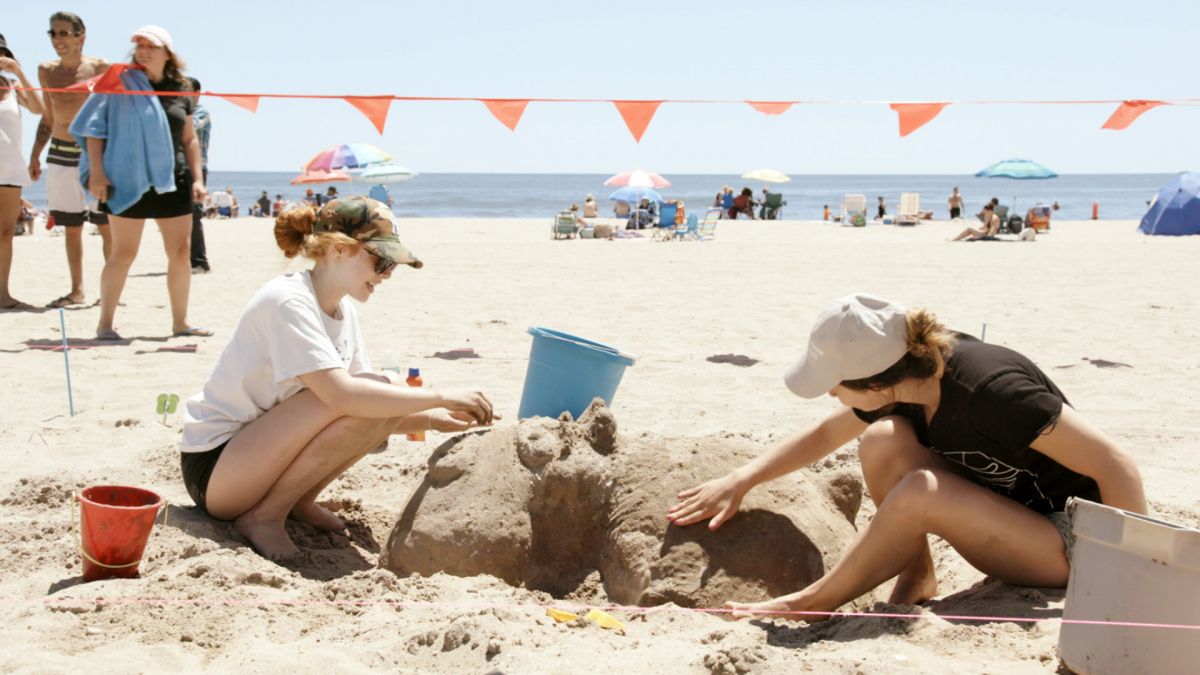 Two women on a busy beach build a sandcastle.