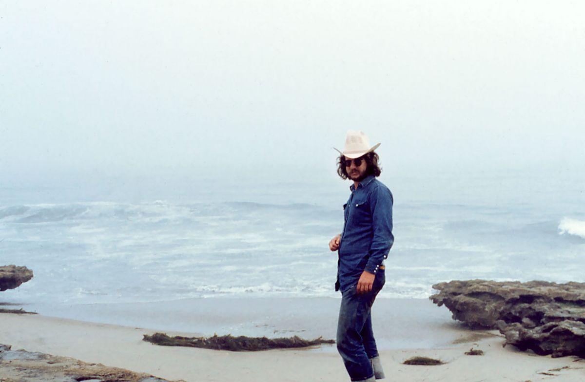 Old photograph of a man with light skin tone standing on the beach. He wears denim shirt and pants and a cowboy hat with sunglasses.