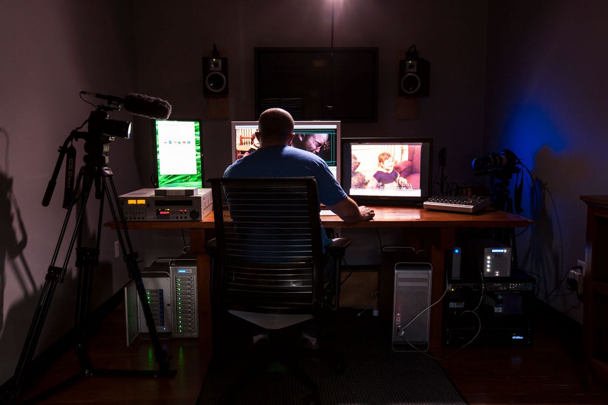 The back of a man in a dimly lit editing suite as he looks over old home movies spread across three computer monitors.