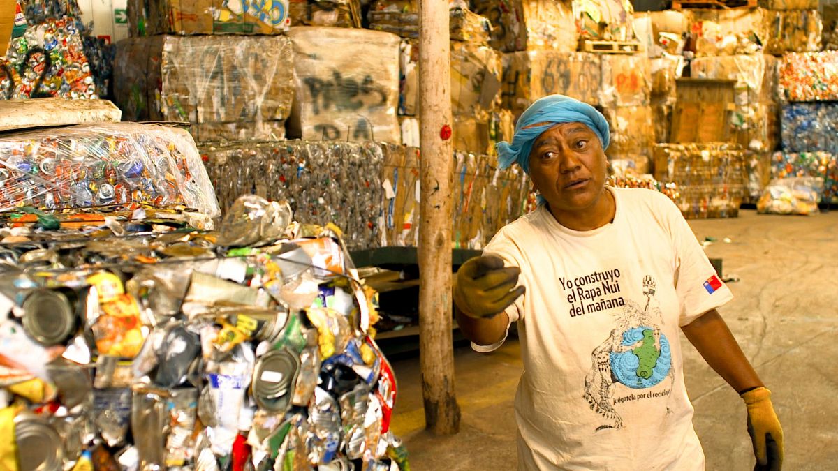 A woman with medium skin tone speaks to the camera while in a recycling facility, with mounds of compressed cans surrounding her.