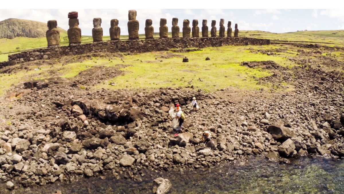 A handful of people clean up a rocky beach on Rapa Nui (Easter Island) while a row of stone moai statues loom in the background.