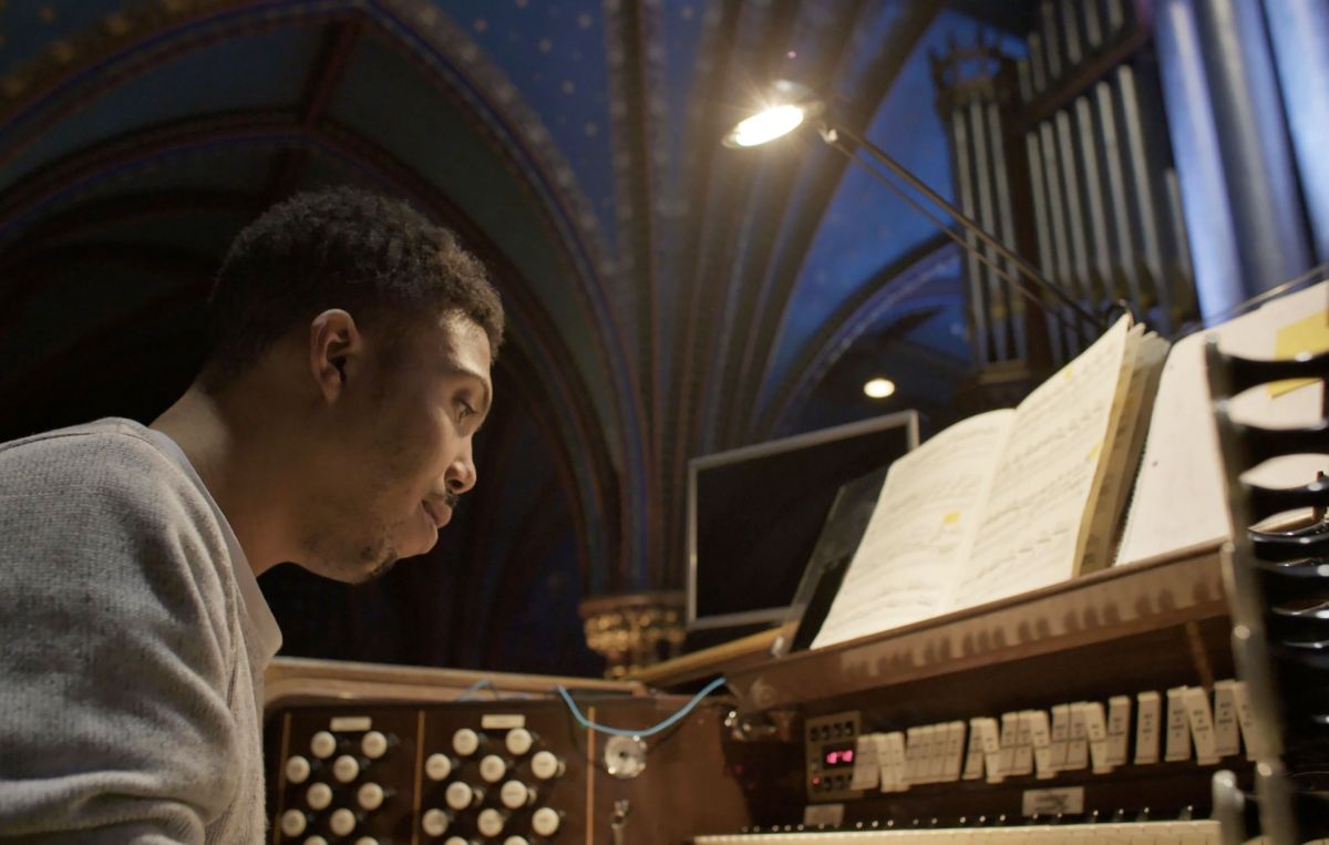 A close-up of a man with dark skin tone as he practices playing a pipe organ in a large church with tall, blue ceilings.