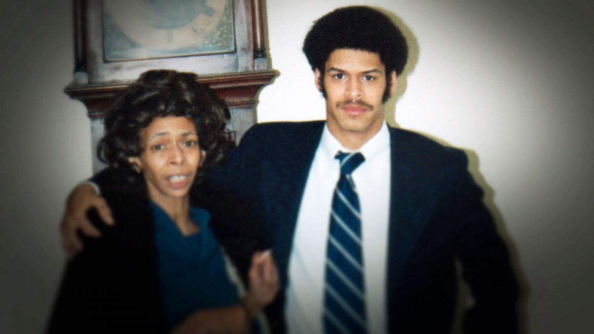 A woman with medium skin tone poses with her son, also with medium skin tone, in a color photograph from the 70s or 80s. They are both dressed formally.